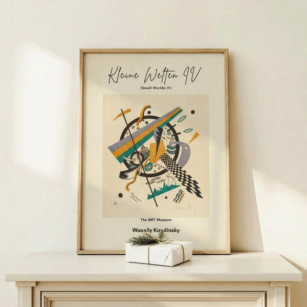 May include: Framed abstract art print in light wood frame displaying geometric shapes, lines, and figures in orange, teal, black, and gray on cream background. Print sits on pale wooden surface against white wall next to small white gift box with greenery sprig.
