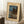 May include: Framed vintage Japanese woodblock print showing a moonlit lakeside scene with traditional buildings, tall dark trees, and mountains under a blue evening sky. The artwork has a cream mat border and thin frame, displayed leaning against a white wall on a textured white rug. A wooden side table with coffee cup and beige throw blanket are visible nearby, creating a cozy home interior setting.
