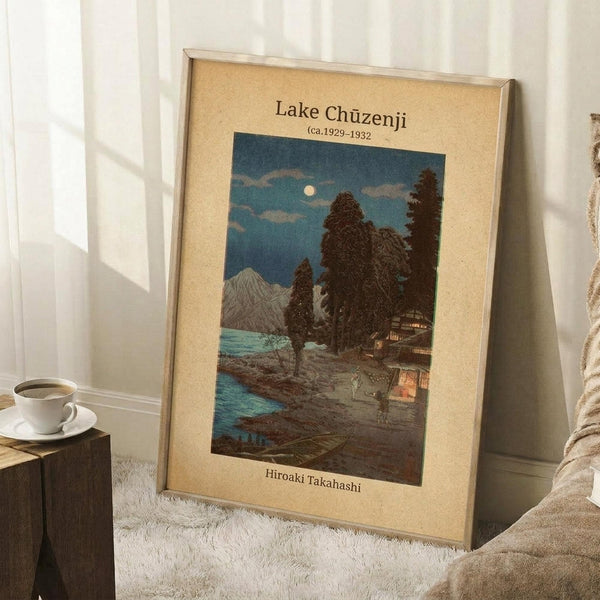 May include: Framed vintage Japanese woodblock print showing a moonlit lakeside scene with traditional buildings, tall dark trees, and mountains under a blue evening sky. The artwork has a cream mat border and thin frame, displayed leaning against a white wall on a textured white rug. A wooden side table with coffee cup and beige throw blanket are visible nearby, creating a cozy home interior setting.