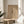 May include: Framed abstract art print with geometric line drawings and text on beige background, displayed on round wooden side table next to white upholstered chair. Natural sunlight creates shadows across the neutral-toned room setting with plant leaves visible in corner.
