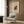 May include: Beige and cream abstract art poster featuring geometric shapes and flowing forms in earth tones, displayed in thin light frame on wood paneled wall above modern cream upholstered armchair with geometric patterned throw pillow, brass floor lamp and dark round side table with open book visible in contemporary neutral-toned room setting.