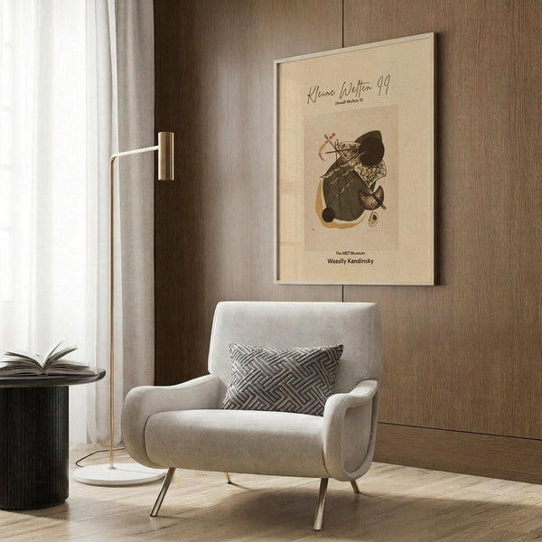 May include: Beige and cream abstract art poster featuring geometric shapes and flowing forms in earth tones, displayed in thin light frame on wood paneled wall above modern cream upholstered armchair with geometric patterned throw pillow, brass floor lamp and dark round side table with open book visible in contemporary neutral-toned room setting.