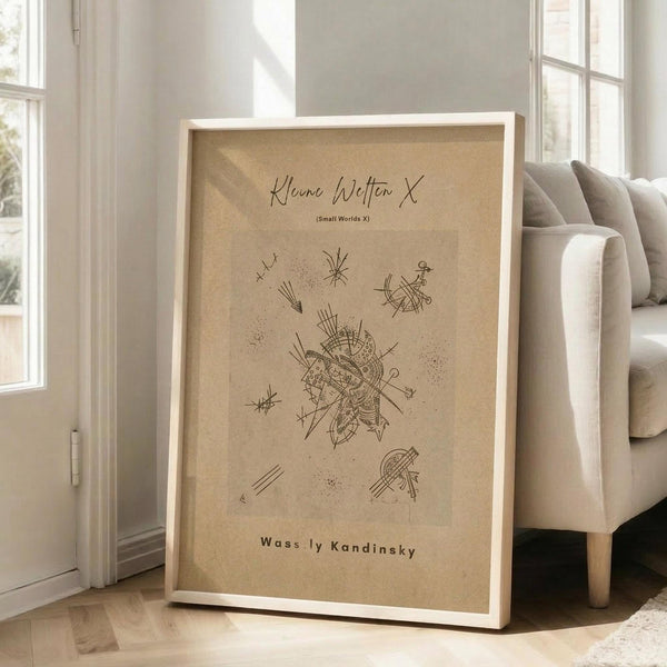 May include: Framed abstract art print with beige background featuring black linear geometric designs and scattered dot patterns, displayed in light wooden frame leaning against white wall next to beige upholstered sofa in bright, sunlit room with white window frames and wooden flooring.