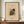 May include: Framed abstract art print in warm wooden frame displayed on walnut credenza alongside white ceramic vase and stacked books. The beige-toned poster features geometric shapes in olive green, gold, and brown with scattered linear elements. Wall has white wainscoting panels. Art appears to be museum exhibition poster style with text at top and bottom.