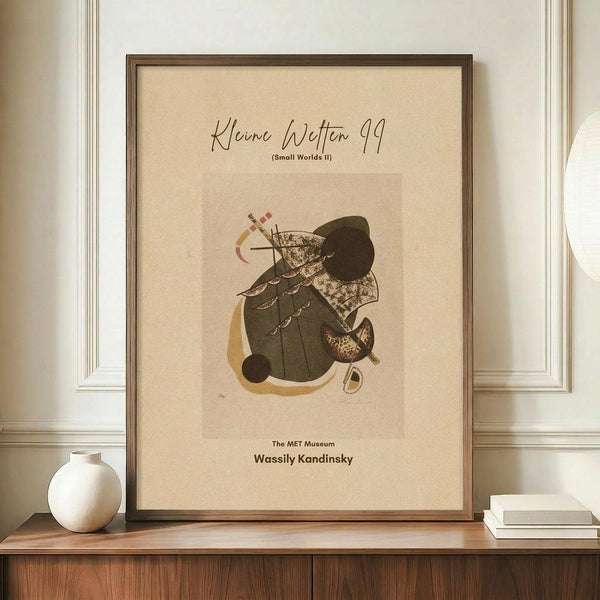 May include: Framed abstract art print in warm wooden frame displayed on walnut credenza alongside white ceramic vase and stacked books. The beige-toned poster features geometric shapes in olive green, gold, and brown with scattered linear elements. Wall has white wainscoting panels. Art appears to be museum exhibition poster style with text at top and bottom.