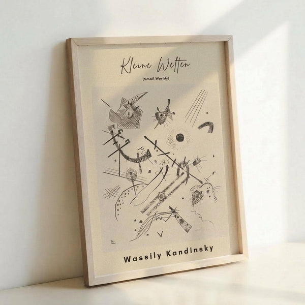 May include: Framed abstract art print in light wooden frame leaning against white wall. Cream-colored poster features black line drawing with geometric shapes, intersecting lines, circles, and angular forms in Kandinsky's distinctive abstract style. Natural lighting creates soft shadows on the minimalist display setup.