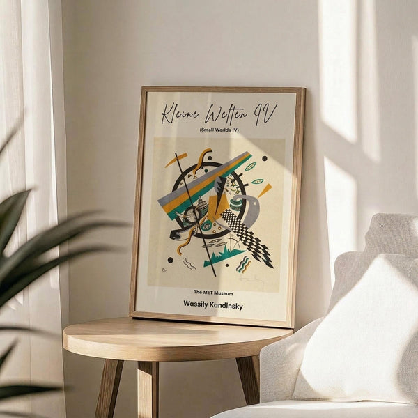May include: Framed abstract art print in thin wood frame displayed on round wooden side table next to beige upholstered chair. Artwork features geometric shapes, lines and forms in orange, teal, black and white with Kandinsky attribution text below. Soft natural lighting creates shadows across white wall background with plant leaves visible at left edge.