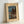 May include: Framed vintage Japanese woodblock print reproduction showing a moonlit lakeside scene with traditional buildings, tall cypress trees, mountains, and turquoise water. The artwork features a cream-colored mat with text identifying the piece and artist. The wooden frame leans against a white wall, displaying the classic blue and earth-toned night landscape in traditional Asian art style.