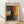May include: Framed abstract art poster leaning against white wall on light wood floor. Features bold blocks of orange, red, yellow, black and gray paint in expressionist style. Natural wood frame with white matting. Soft window light creates shadows on textured wall surface.