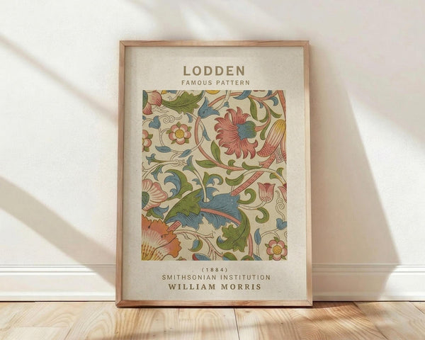 May include: Framed botanical print featuring William Morris's ornate floral pattern in muted greens, coral pink, and blue tones against cream background. The decorative design shows flowing leaves, stylized flowers, and curving vines in Arts and Crafts style. Natural wood frame sits on light wooden floor against white wall with soft shadows from natural lighting.