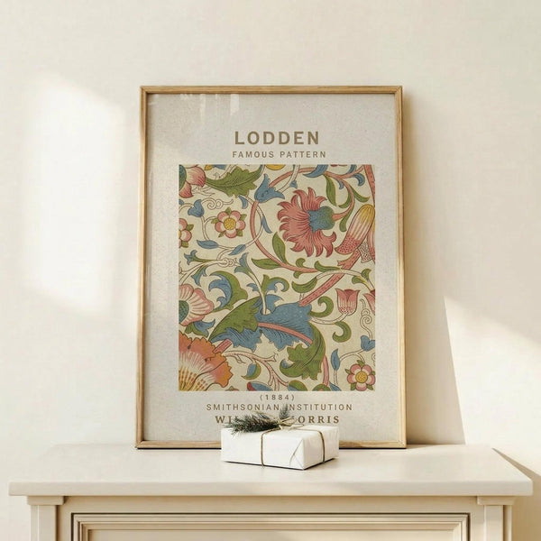 May include: Framed botanical print featuring William Morris floral pattern with intricate leaves, flowers and vines in coral pink, sage green, and dusty blue colors on cream background. Natural wood frame displayed on light wooden surface next to small white wrapped gift with evergreen sprig. Neutral beige wall backdrop with soft natural lighting creating gentle shadows.