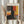 May include: Hands holding a framed abstract expressionist print featuring bold blocks of orange, red, yellow, and dark colors with painterly brushstrokes. Light wooden frame against white curtained window with dried pampas grass visible to the side. Person wearing cream colored sweater with gathered cuffs.
