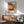 May include: Framed Mark Rothko exhibition poster with orange and brown abstract artwork hanging above wooden sideboard. Modern living room setting features mint green upholstered armchair with wooden arms, white ceramic table lamp, and round coffee table. Warm natural lighting creates shadows on white wall. Mid-century modern furniture styling with clean lines and natural wood tones.