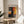 May include: Abstract expressionist artwork poster in thin wooden frame displayed on round wooden side table next to beige upholstered chair. The artwork features bold vertical blocks of yellow, orange, red and dark colors with soft edges. Natural sunlight streams across white walls creating geometric shadows, with green plant leaves visible in corner of bright, minimalist room setting.
