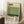 May include: Framed vintage-style art poster with cream background featuring horizontal color blocks in yellow, sage green, and dark green. Bold black text reads "Mark Rothko" at top and birth/death dates at bottom right. Wooden frame leans against white wall next to white coffee cup on wooden surface and textured cream throw blanket on white fuzzy rug.
