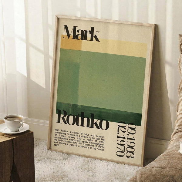 May include: Framed vintage-style art poster with cream background featuring horizontal color blocks in yellow, sage green, and dark green. Bold black text reads "Mark Rothko" at top and birth/death dates at bottom right. Wooden frame leans against white wall next to white coffee cup on wooden surface and textured cream throw blanket on white fuzzy rug.