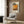 May include: Modern living room with beige armchair featuring geometric patterned pillow, gold floor lamp, dark round side table with open book, and large framed art poster on wood-paneled wall. The poster displays Mark Rothko's name with orange and brown abstract color field artwork and biographical text, creating a sophisticated mid-century modern interior setting with warm neutral tones.