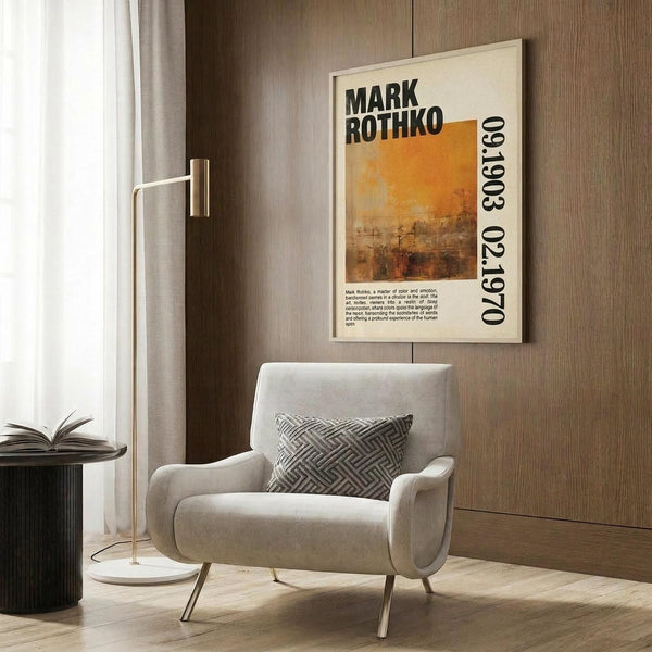 May include: Modern living room with beige armchair featuring geometric patterned pillow, gold floor lamp, dark round side table with open book, and large framed art poster on wood-paneled wall. The poster displays Mark Rothko's name with orange and brown abstract color field artwork and biographical text, creating a sophisticated mid-century modern interior setting with warm neutral tones.