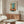 May include: Abstract art poster with teal, orange, and cream color blocks in the style of Mark Rothko, displayed in thin wooden frame above natural wood credenza. Modern living room setting with white curved chair, glass oval coffee table, and table lamp. Minimalist Scandinavian-style interior with neutral walls and natural lighting.