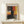 May include: Abstract expressionist artwork poster in wooden frame displayed on light wood console table against white wall. Print features bold blocks of orange, red, yellow, black and gray paint with brushy, layered texture. Small white wrapped gift with twine and greenery sits beside frame. Warm, minimalist home decor setting with natural lighting.
