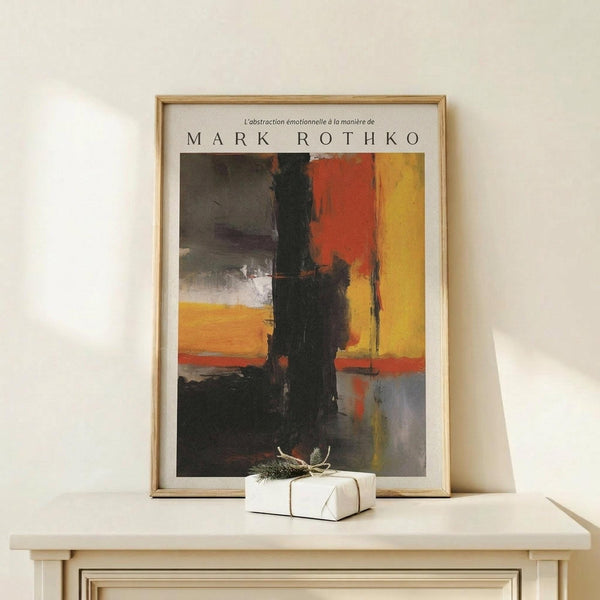 May include: Abstract expressionist artwork poster in wooden frame displayed on light wood console table against white wall. Print features bold blocks of orange, red, yellow, black and gray paint with brushy, layered texture. Small white wrapped gift with twine and greenery sits beside frame. Warm, minimalist home decor setting with natural lighting.