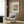 May include: Framed William Morris botanical pattern print with colorful floral design hanging on wood-paneled wall above beige upholstered armchair with geometric pillow. Gold floor lamp and dark side table with open book visible. Modern interior setting with warm wood tones and neutral textiles.
