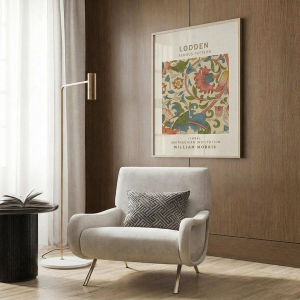 May include: Framed William Morris botanical pattern print with colorful floral design hanging on wood-paneled wall above beige upholstered armchair with geometric pillow. Gold floor lamp and dark side table with open book visible. Modern interior setting with warm wood tones and neutral textiles.