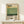 May include: Framed poster with Mark Rothko's name in bold black letters above abstract color blocks in cream, yellow, and varying shades of green. Text below describes the artist with birth and death dates. Natural wood frame sits on light wooden surface next to small white wrapped gift with twine and greenery sprig against cream wall.