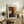 May include: Framed abstract art print in light wood frame displayed on rustic wooden sideboard alongside fern plant, ceramic vase, and books. Poster features bold blocks of orange, red, yellow and dark colors in painterly style. Neutral living room setting with beige chair and warm lighting.