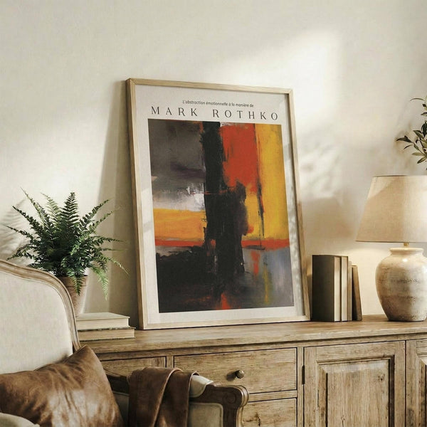 May include: Framed abstract art print in light wood frame displayed on rustic wooden sideboard alongside fern plant, ceramic vase, and books. Poster features bold blocks of orange, red, yellow and dark colors in painterly style. Neutral living room setting with beige chair and warm lighting.