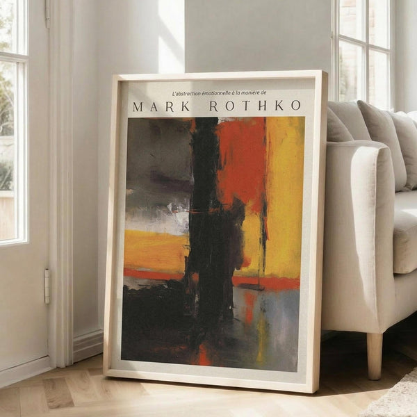 May include: Large framed abstract art print leaning against white wall in bright living room. Artwork features bold vertical brushstrokes in yellow, orange, red, and black with "MARK ROTHKO" text at top. Light wood frame, positioned next to beige upholstered sofa on hardwood floors with natural sunlight streaming through tall windows.