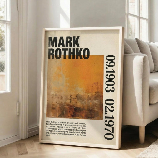 May include: Framed art poster with Mark Rothko's name in bold black letters above an abstract painting featuring warm orange and brown gradients. White matted frame leans against light gray wall next to beige sofa with pillows. Natural sunlight streams through tall windows onto hardwood floors with area rug. The poster includes biographical text about the artist and shows weathered, textured brushwork in earthy tones.