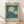 May include: Framed botanical poster print leaning against white wall on light wood floor. Features two white daisy flowers with yellow centers on teal blue background with dark green leaves. Text reads "FLOWER MARKET" at top and "LISBON No.4 Gallery Prints Color Botanical Garden of lisbon" at bottom on cream colored border. Natural wood frame with soft shadows from window light on textured white wall.