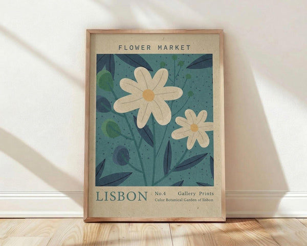 May include: Framed botanical poster print leaning against white wall on light wood floor. Features two white daisy flowers with yellow centers on teal blue background with dark green leaves. Text reads "FLOWER MARKET" at top and "LISBON No.4 Gallery Prints Color Botanical Garden of lisbon" at bottom on cream colored border. Natural wood frame with soft shadows from window light on textured white wall.