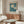 May include: Framed botanical poster with white daisies on teal green background displayed above wooden sideboard in modern living room with cream chair, glass coffee table, and white table lamp