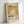 May include: Framed art print leaning against white wall featuring abstract golden yellow and cream colored blocks with "Mark Rothko" text and "02.1970" dates. Print has vintage-inspired typography and descriptive text about the artist. Light wood frame with beige matting, photographed in natural lighting showing shadows on wall.