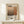 May include: Framed art poster featuring Mark Rothko's name in large black text above abstract geometric shapes in warm browns and tans, with biographical text and dates 1903-1970. Light wood frame displayed on white wooden console table against cream wall, with small wrapped gift box with greenery beside it.
