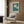 May include: Vintage-style botanical poster featuring white daisies on teal background with "Flower Market" and "Lisbon" text, displayed in light wood frame above beige upholstered armchair with geometric patterned pillow. Modern brass floor lamp and black side table with open book complete the neutral-toned living room setting with wood paneled walls.