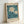 May include: Framed botanical poster print leaning against white wall, featuring two white daisies with yellow centers on teal blue background with dark green leaves and stems, vintage-style design with "FLOWER MARKET" and "LISBON" text at top and bottom, displayed in light wood frame on neutral surface