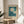 May include: Framed botanical art print displayed on round wooden side table next to beige upholstered chair. Print features two white daisies with yellow centers against teal blue background with dark green leaves. Text reads "Flower Market" and "Lisbon" on cream-colored poster. Natural wood frame catches warm sunlight streaming through window, creating soft shadows on neutral wall.
