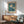 May include: Framed botanical poster featuring white daisies with yellow centers against teal blue background with green leaves, displayed above wooden sideboard with ceramic lamp in modern living room setting. Mid-century style chair with sage green upholstery and wooden arms visible in corner. Natural sunlight creates shadows on white wall.