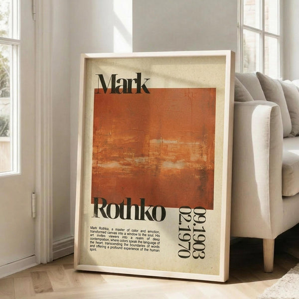 May include: Framed vintage-style poster with cream and orange color blocks, featuring "Mark Rothko" text and biographical description. White wooden frame leaning against white wall corner next to beige upholstered sofa with pillows. Bright natural light streams through tall windows onto hardwood floors with area rug in minimalist living room setting.