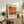 May include: Framed poster featuring Mark Rothko's name and orange abstract painting reproduction displayed on rustic wooden sideboard. Room setting shows neutral walls, fern plant, ceramic vase, stacked books, and beige upholstered chair with brown throw blanket. Natural lighting creates soft shadows across the vintage-style interior decor.