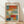 May include: Framed abstract art print in light wood frame leaning against white wall on wooden floor. Artwork features soft rectangular blocks of color including orange, red, blue, yellow, and white with painterly, blended edges in style reminiscent of Mark Rothko. Natural lighting creates diagonal shadows across the scene. Frame appears medium-sized relative to baseboard and flooring.
