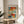 May include: Abstract colorful poster with geometric blocks in blues, oranges, reds, and yellows displayed in thin gold frame on round wooden side table next to beige upholstered chair, with plant shadows cast on white wall from natural sunlight
