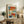 May include: Abstract colorful poster print with blue, orange, red, and yellow rectangular blocks in wooden frame, displayed on rustic wood sideboard next to fern plant, ceramic vase, stacked books, and beige upholstered chair in neutral-toned living room setting