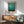 May include: Framed teal and turquoise abstract art print hanging on white wall above wooden sideboard with white table lamp. Light green upholstered armchair with wooden frame sits nearby on wooden floor with natural sunlight casting shadows across the minimalist room setting.