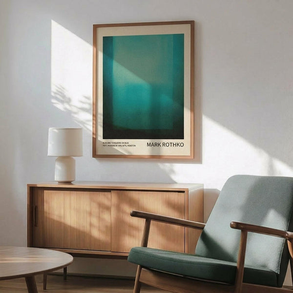 May include: Framed teal and turquoise abstract art print hanging on white wall above wooden sideboard with white table lamp. Light green upholstered armchair with wooden frame sits nearby on wooden floor with natural sunlight casting shadows across the minimalist room setting.