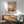 May include: Framed abstract art print with warm orange, pink and brown tones in wooden frame, displayed above light wood sideboard with white table lamp. Mid-century modern green upholstered armchair with wooden arms sits nearby. Bright natural light creates geometric shadows on white wall in minimalist living room setting.