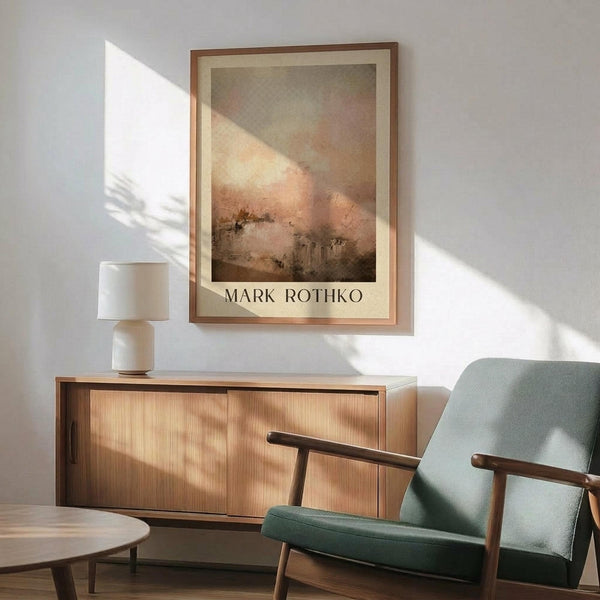 May include: Framed abstract art print with warm orange, pink and brown tones in wooden frame, displayed above light wood sideboard with white table lamp. Mid-century modern green upholstered armchair with wooden arms sits nearby. Bright natural light creates geometric shadows on white wall in minimalist living room setting.